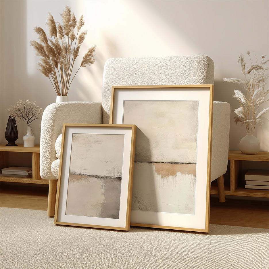 Two framed Scandinavian Modern Neutral Abstract Wall Art - Beige Minimalist Canvas Print pieces with soft beige and gray tones leaning against a textured white armchair in a warm, cozy living room setting with natural light and dried pampas grass accents.