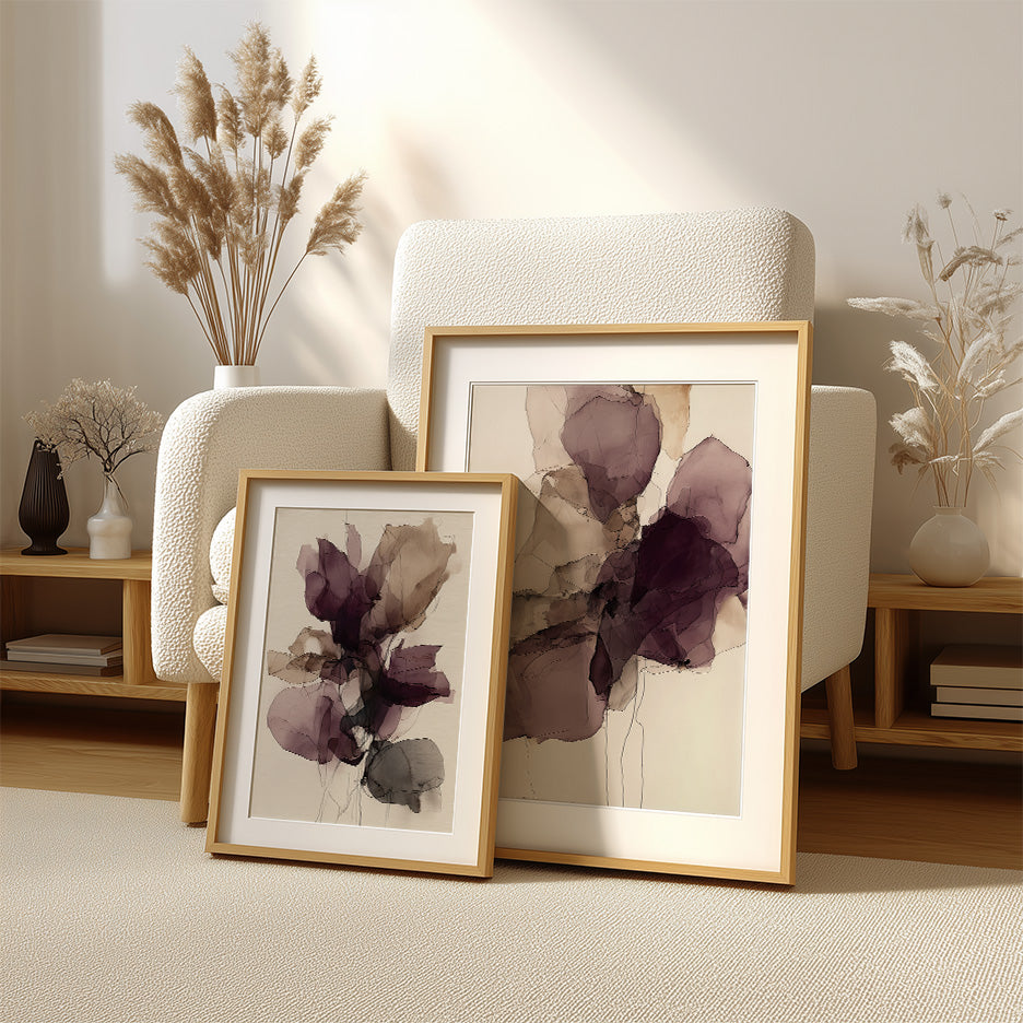 Two framed ink wash abstract floral wall canvas art prints in burgundy tones leaning against a textured cream armchair in a minimalist modern living room setting, featuring natural light and neutral decor accents, showcasing the Ink Wash Abstract Floral Wall Canvas Art Print - Burgundy Minimalist Modern Wall Art.