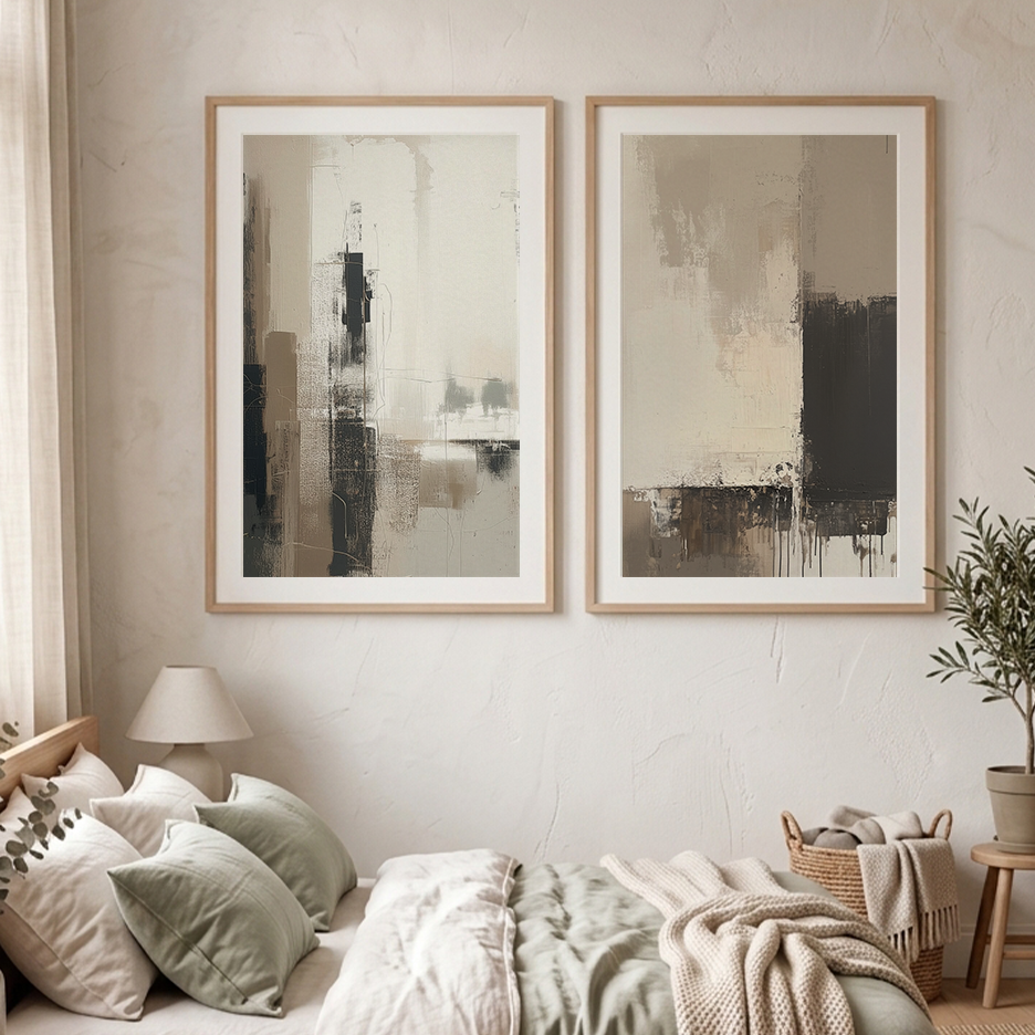 Two framed abstract wall art prints in light wood frames hang above a bed in a softly styled bedroom. The artwork features neutral tones of beige, gray, brown, and black, and the room includes a lamp, pillows, blanket, and a potted plant.