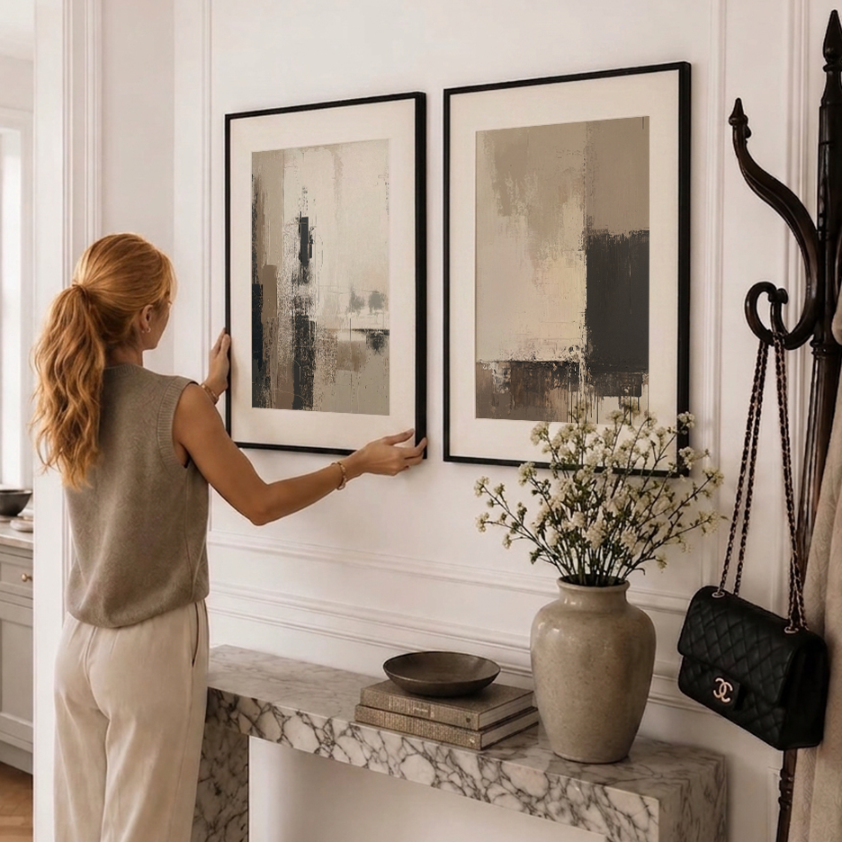 A woman in a beige sleeveless top and cream pants hangs two framed abstract art prints on a white paneled wall in a styled entryway. The prints have black frames and a neutral color palette of beige, gray, and black.