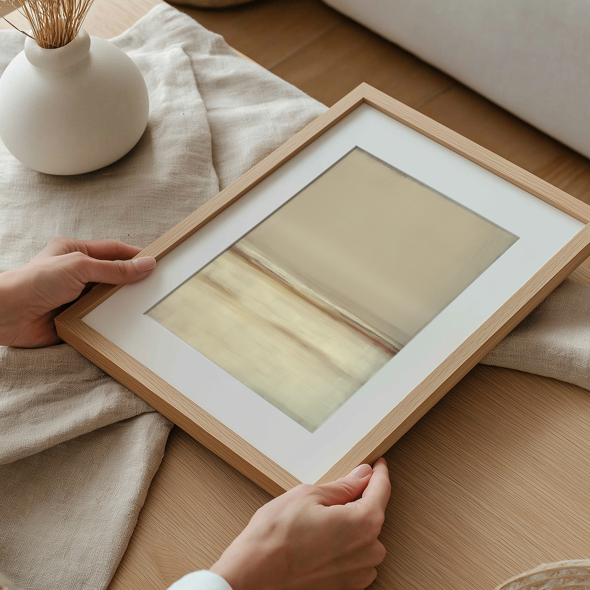 Hands holding a framed Neutral Seascape Wall Art | Beige Abstract Canvas with soft beige and cream tones, set on a light wooden table with a white vase nearby.