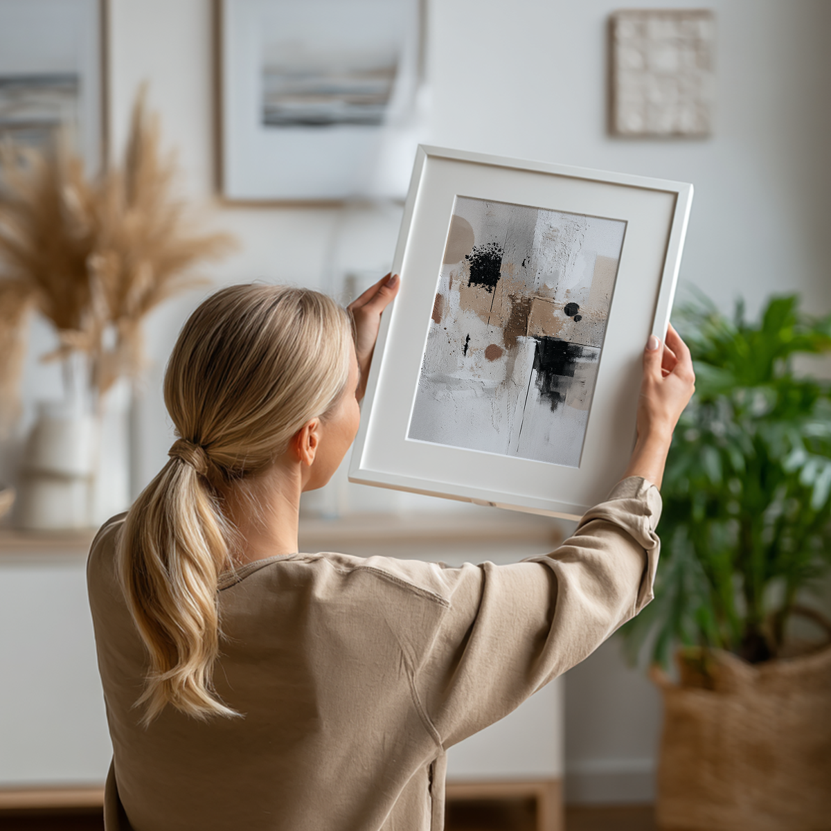 Woman holding a framed neutral textured abstract wall art with beige, black, and white tones in a bright, minimalist living room setting, featuring the Neutral Textured Abstract Wall Art | Japandi Minimalist Canvas.