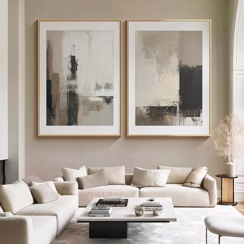 Two large framed abstract wall art prints in beige, gray, and black tones hang above a cream sofa in a modern living room setting.