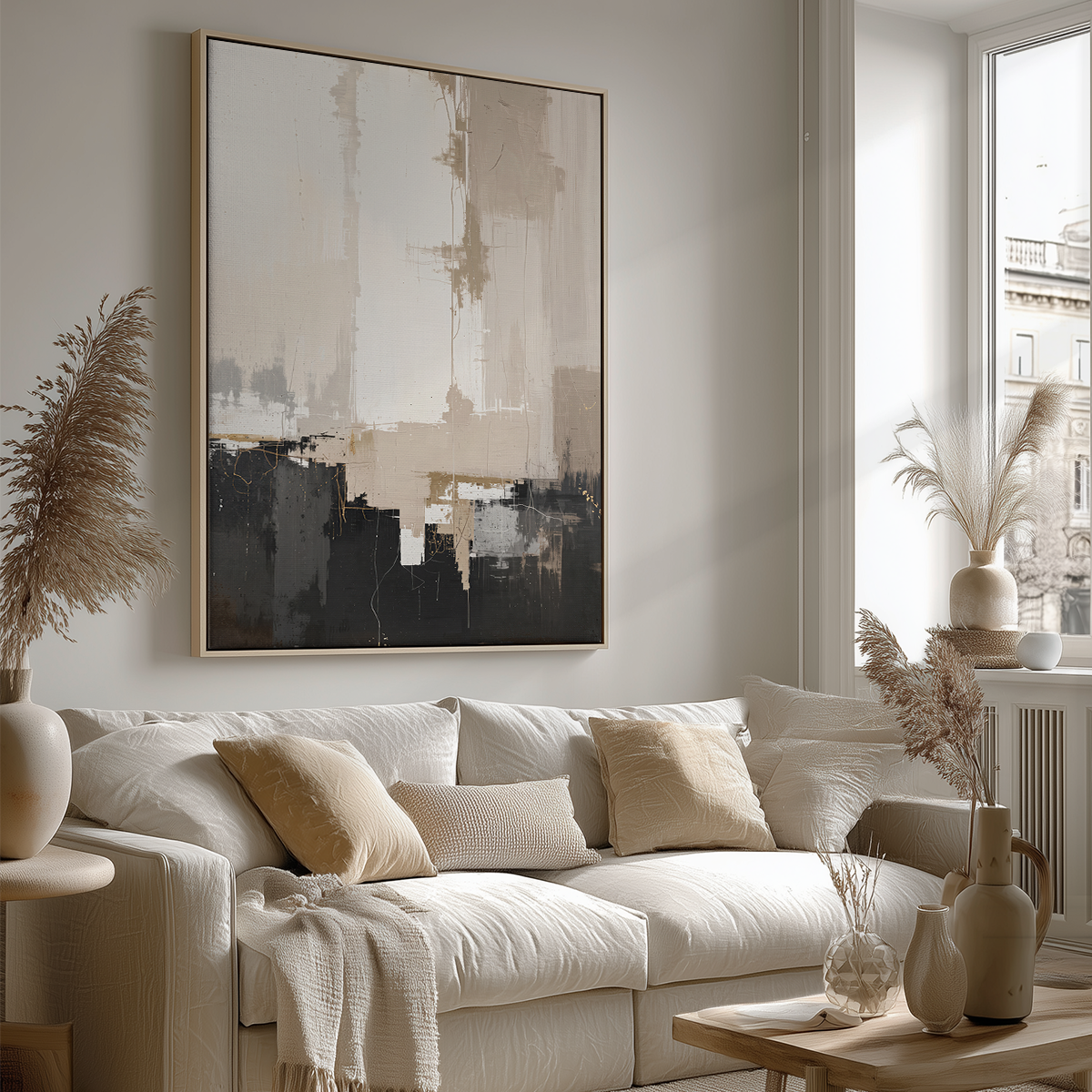 A large framed abstract wall art print hangs above a cream-colored sofa in a bright, modern living room. The artwork features a minimalist design with beige, gray, and black brushstrokes, and the room is styled with neutral-toned pillows, vases, and dried grasses.