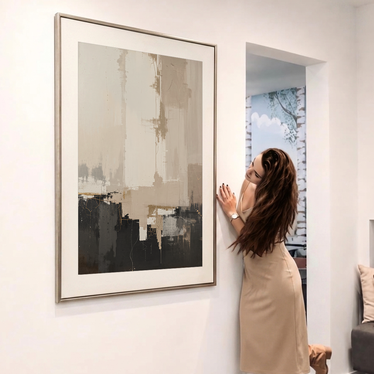 A woman in a beige dress leans against a white wall next to a large framed abstract painting. The painting features a modern abstract design with beige, gray, and black brushstrokes, set in a thin metallic frame.