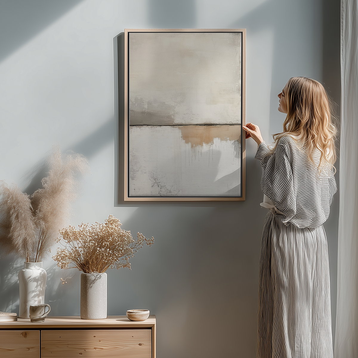 A woman in a striped dress admiring a Scandinavian Modern Neutral Abstract Wall Art - Beige Minimalist Canvas Print hanging on a light gray wall above a wooden sideboard with dried flowers and decor.