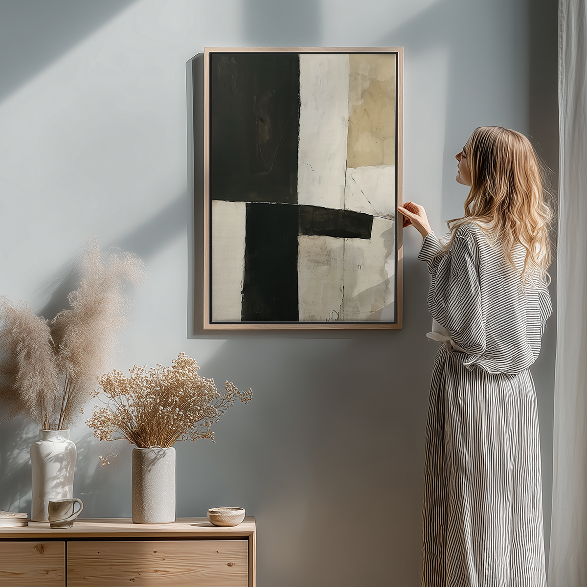 Woman admiring Geometric Abstract Wall Art | Neutral Black Beige Minimalist Print | Modern Wabi-Sabi Decor hanging on a light gray wall above a wooden sideboard with dried floral arrangements in a minimalist room.
