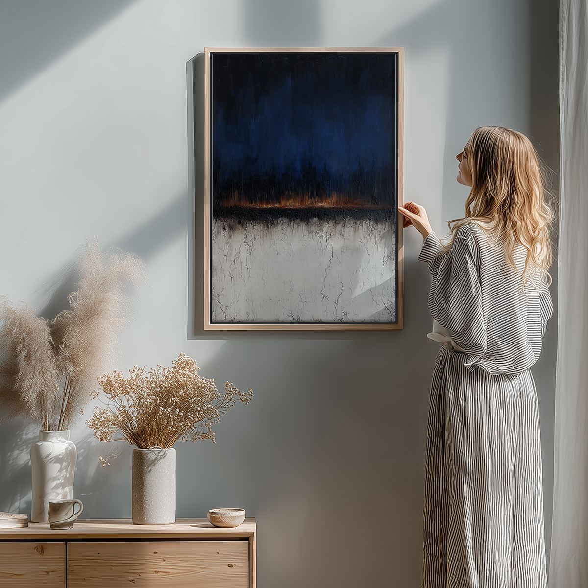 Woman in striped dress adjusting Moody Horizon Abstract Wall Art | Dark Minimalist Landscape Print | Navy & Burnt Orange Decor on a light gray wall above a wooden cabinet with dried plants.
