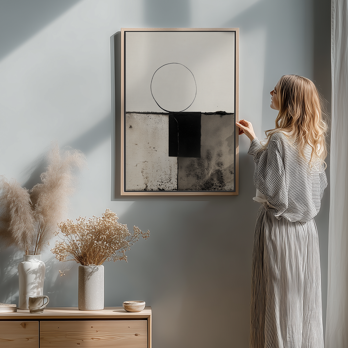 A woman in a striped dress admiring a Modern Abstract Geometric Wall Art – Minimalist Black & White Textured Print hanging on a light-colored wall above a wooden cabinet with neutral decor.