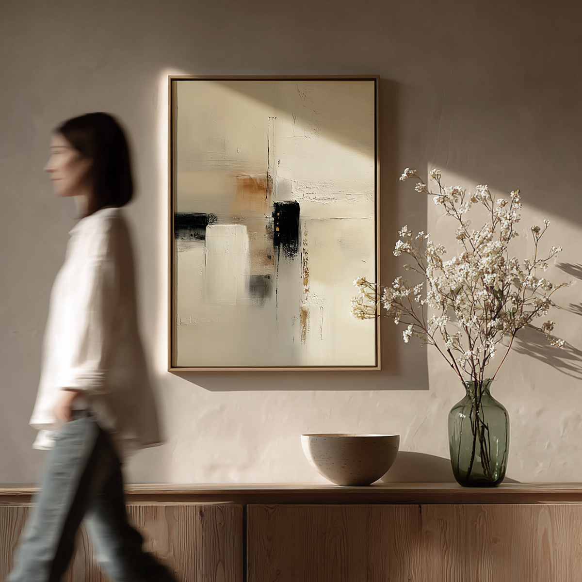 Japandi Abstract Wall Art | Neutral Textured Canvas with Gold Accents | Modern Minimalist Decor hanging on a muted beige wall above a wooden sideboard with a ceramic bowl and a green glass vase holding delicate white flowers, with a blurred woman walking past.