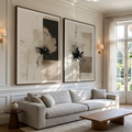 A styled living room featuring two large framed abstract floral wall art prints in black, white, and beige tones, hung above a light beige sofa. The room is decorated with warm wall sconces, a wooden coffee table, and a window with sheer curtains.