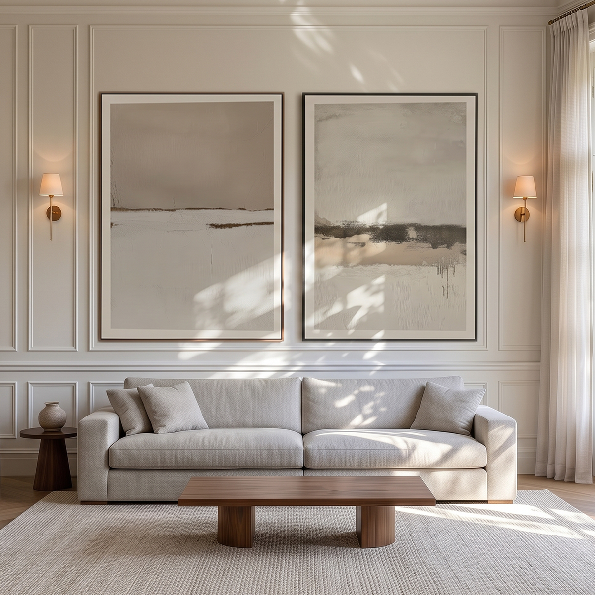 A styled living room featuring two large, minimalist abstract canvas prints in beige and gray tones, hung on a cream paneled wall above a light beige sofa with matching pillows, a wooden coffee table, and a textured cream rug.